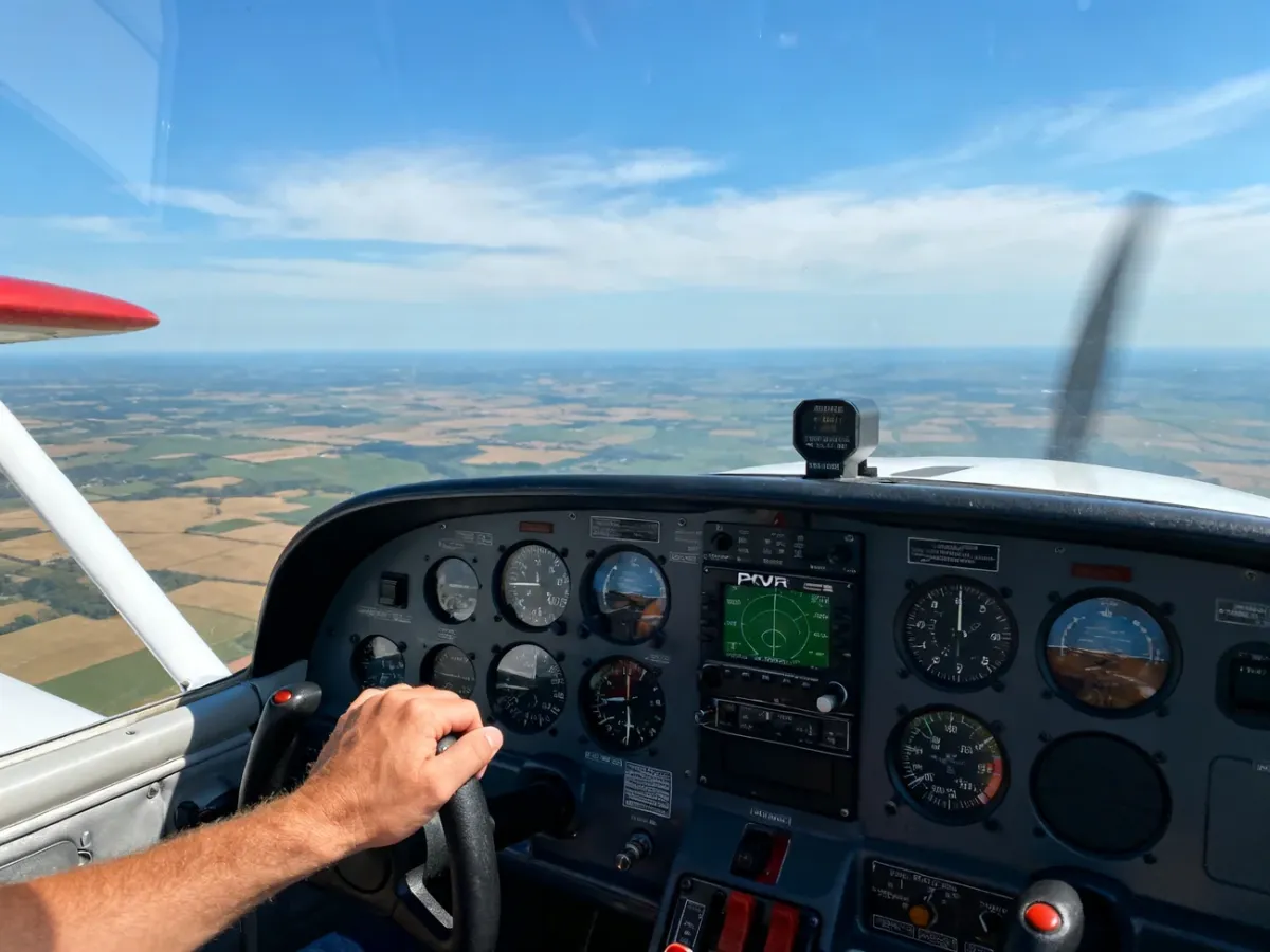 Widok kokpitu małego samolotu w locie z widocznymi przyrządami VOR, pilot trzyma drążek, w tle krajobraz pól i nieba.